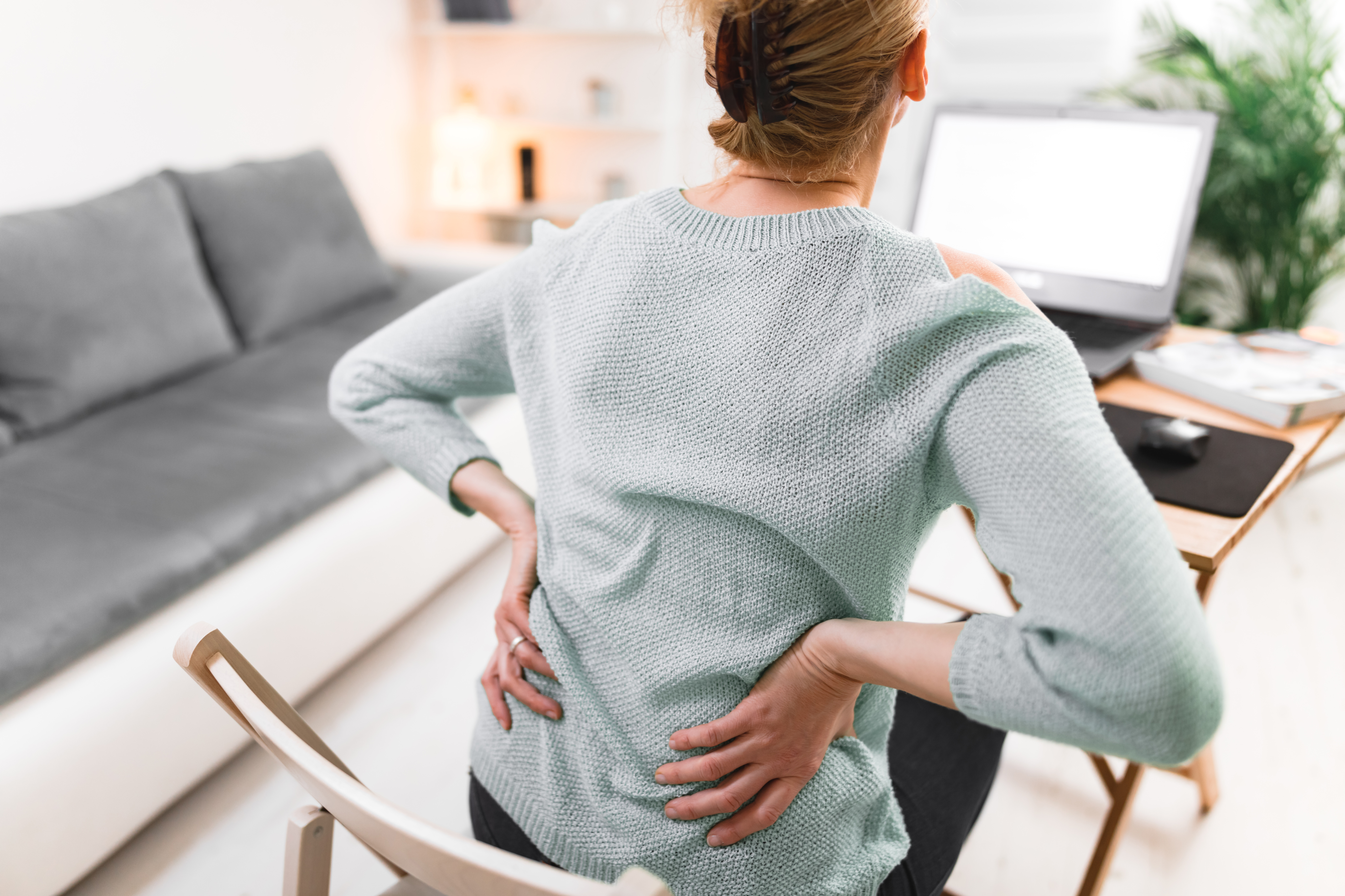 A woman experiencing lower back pain muscle problems as she sits in a chair, working on a computer lap top at a desk.