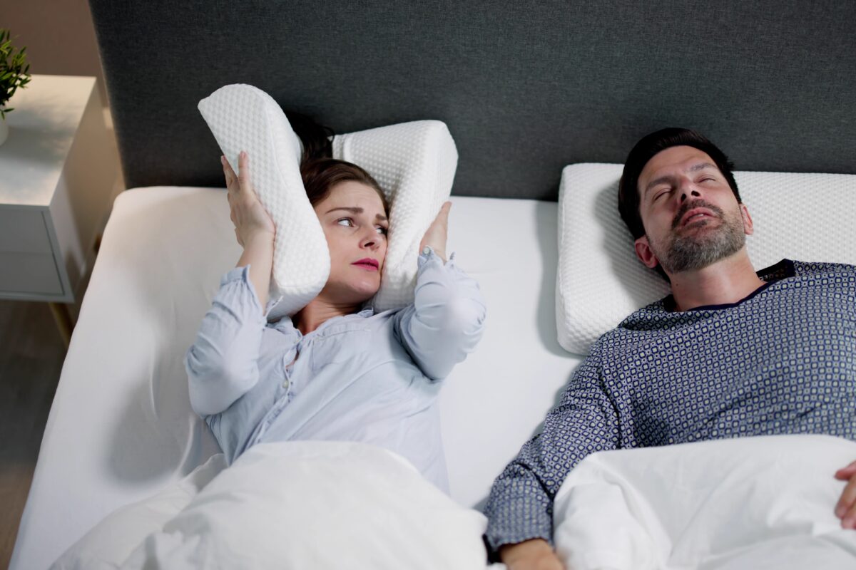 OSA treatment can help men, as illustrated by this married couple in bed as the wife covers her ears with a pillow due to her spouse's loud sleep apnea snoring.