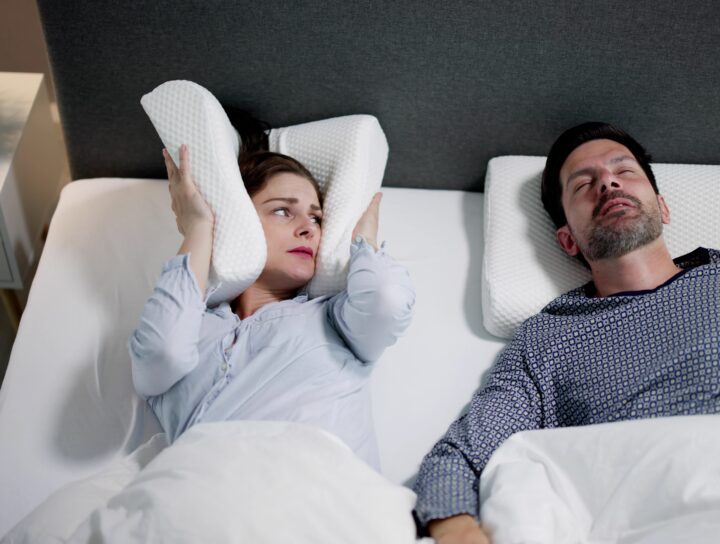 OSA treatment can help men, as illustrated by this married couple in bed as the wife covers her ears with a pillow due to her spouse's loud sleep apnea snoring.