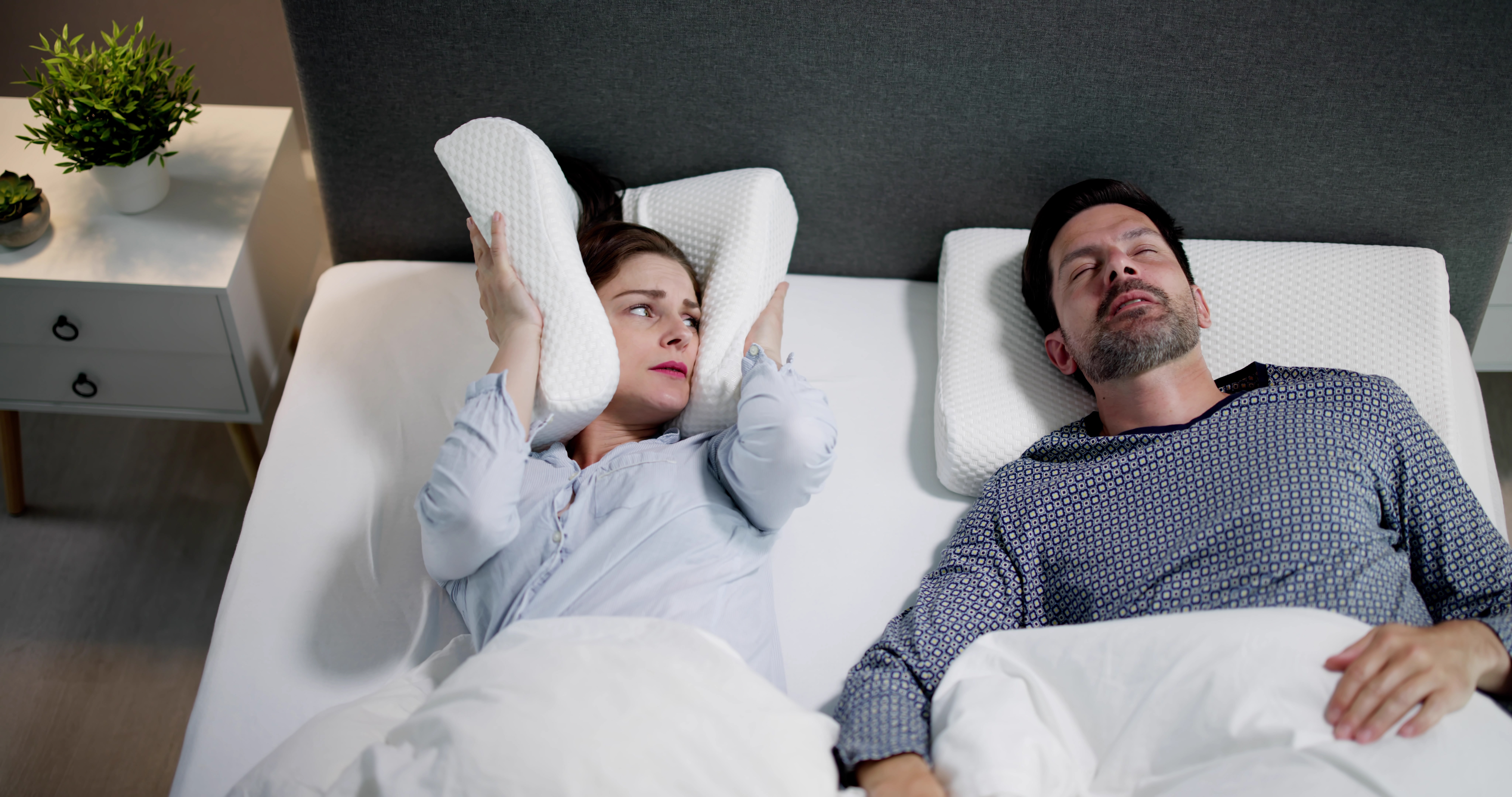 OSA treatment can help men, as illustrated by this married couple in bed as the wife covers her ears with a pillow due to her spouse's loud sleep apnea snoring.