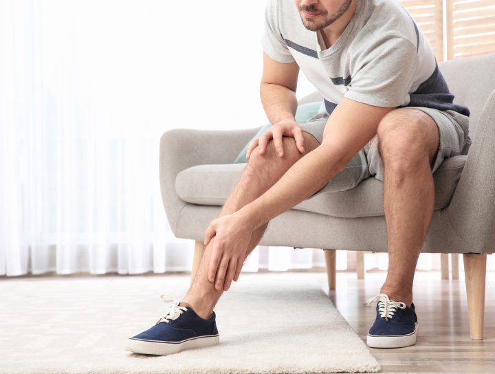 A man sitting on his couch, dealing with painful lower legs and massaging one of them.