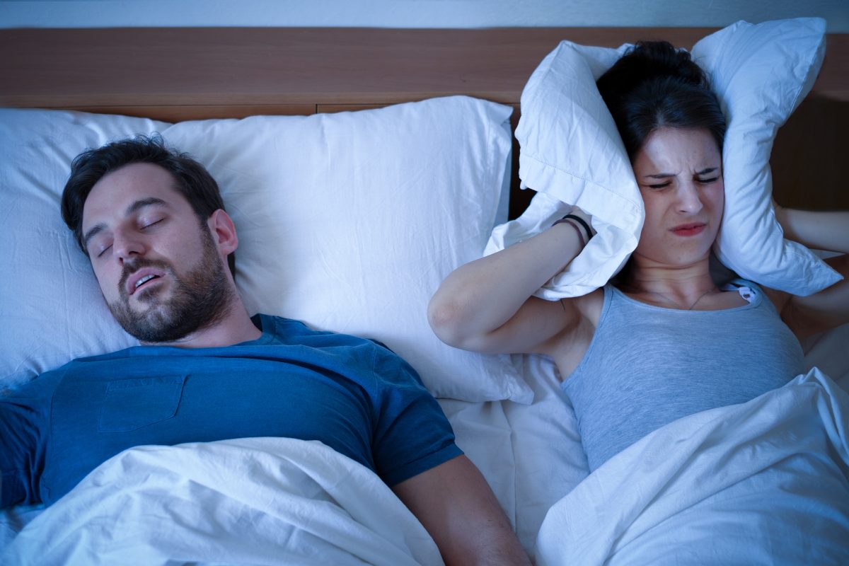 A woman covers her ears in the middle of the night in bed, considering CPAP alternatives as she sleeps next to her snoring husband.