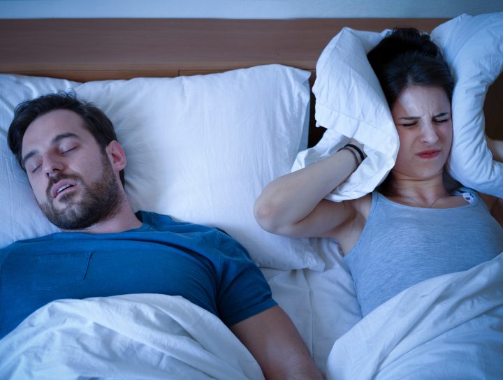 A woman covers her ears in the middle of the night in bed, considering CPAP alternatives as she sleeps next to her snoring husband.
