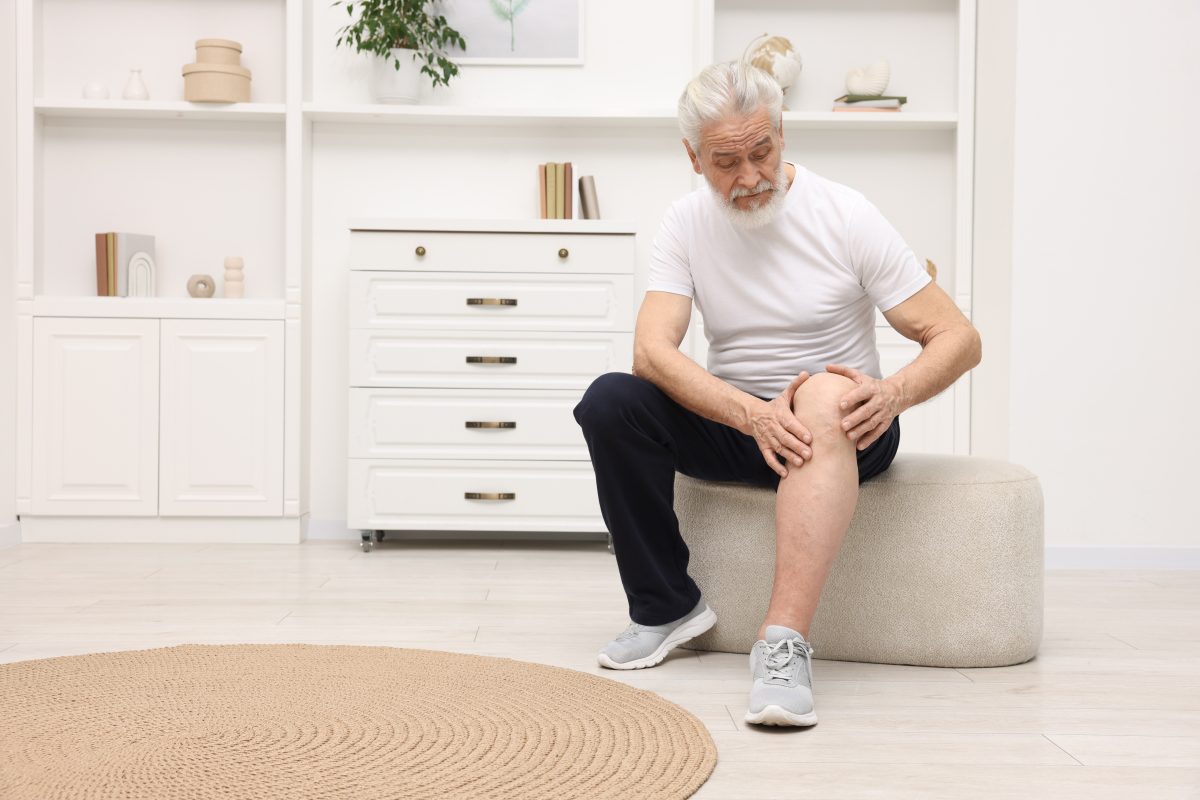 A senior man sitting down and examining his knee with his pant-leg rolled up, considering knee pain treatment.