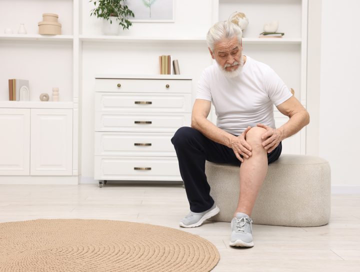 A senior man sitting down and examining his knee with his pant-leg rolled up, considering knee pain treatment.