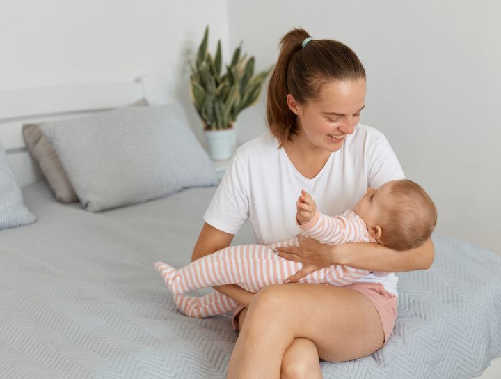 A mother wearing shorts and sitting on the end of her bed smiles at her baby as she holds him, crossing her legs and thinking about spider vein removal.
