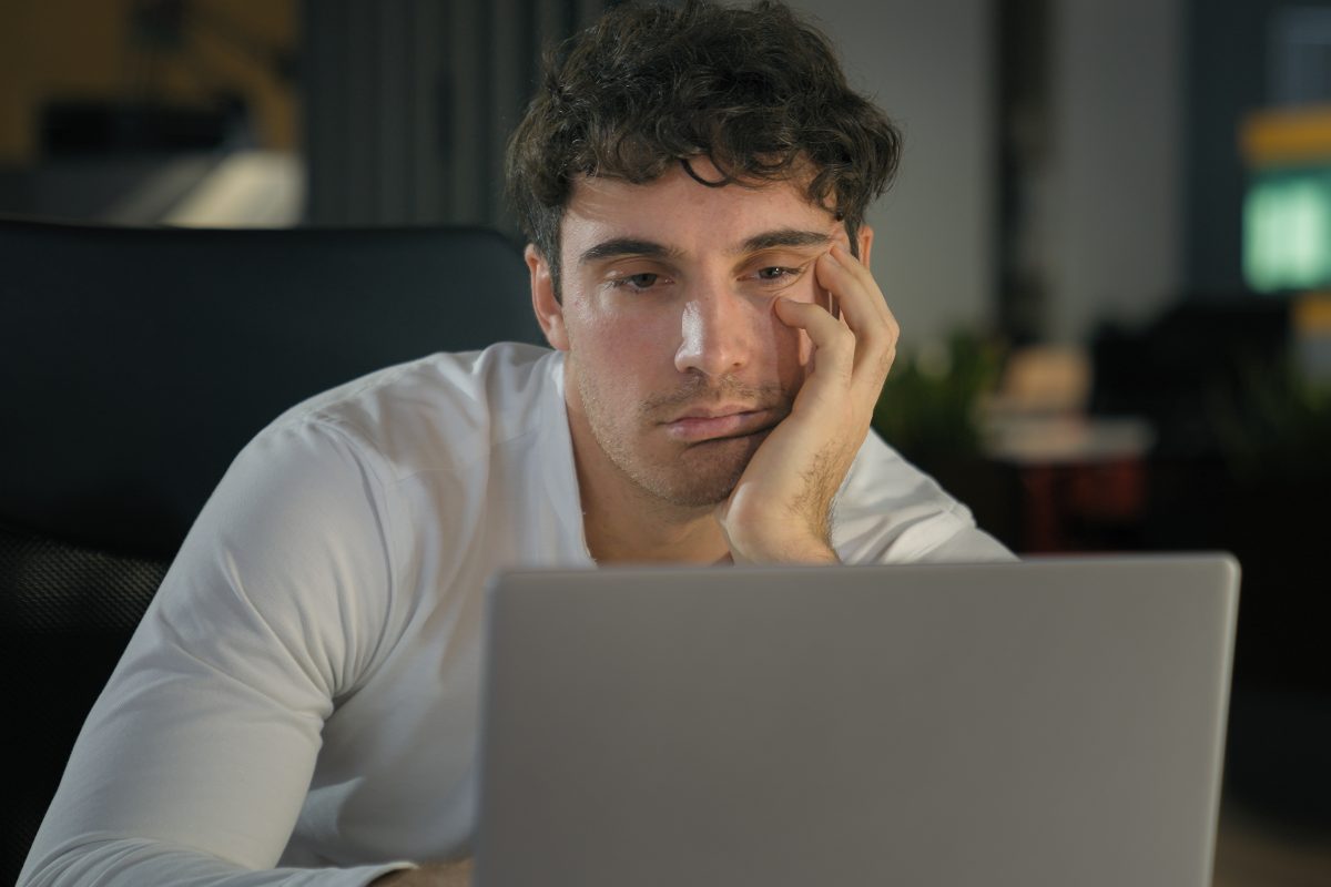 A tired man sitting in front of his lap top and working, considering the benefits of a home sleep test.