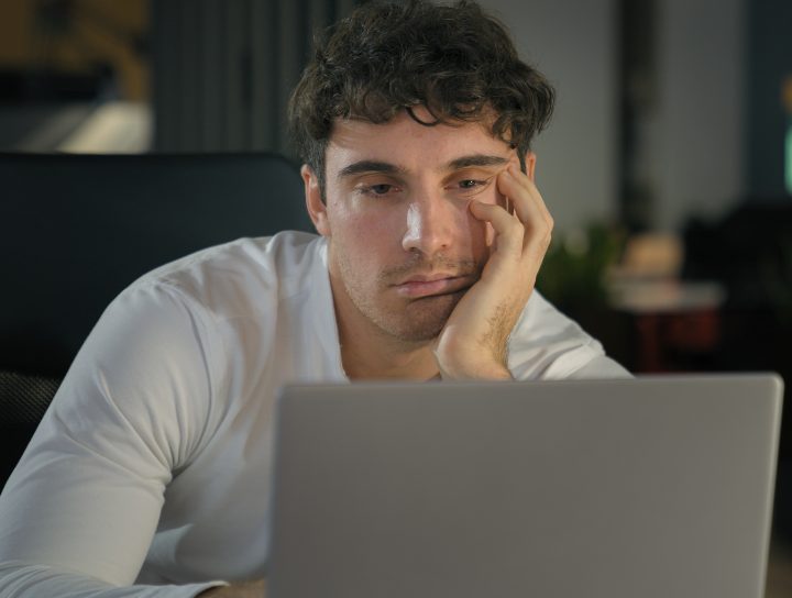 A tired man sitting in front of his lap top and working, considering the benefits of a home sleep test.